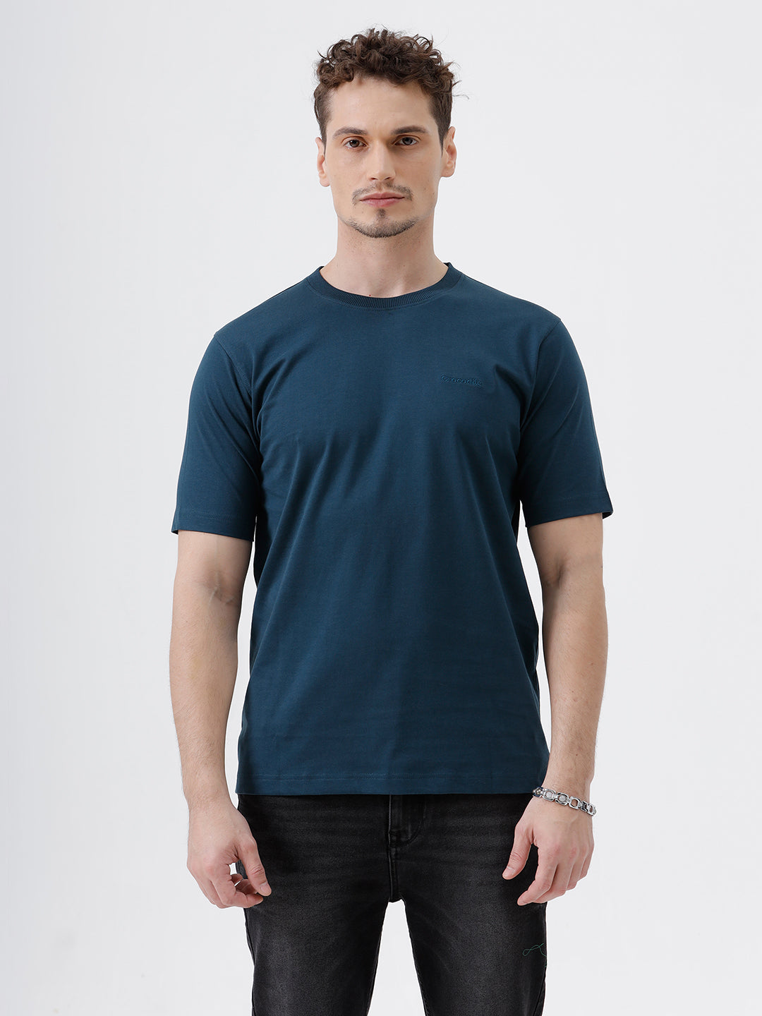 A man wearing a dark navy blue crew-neck t-shirt and black jeans, standing against a light gray studio background.
