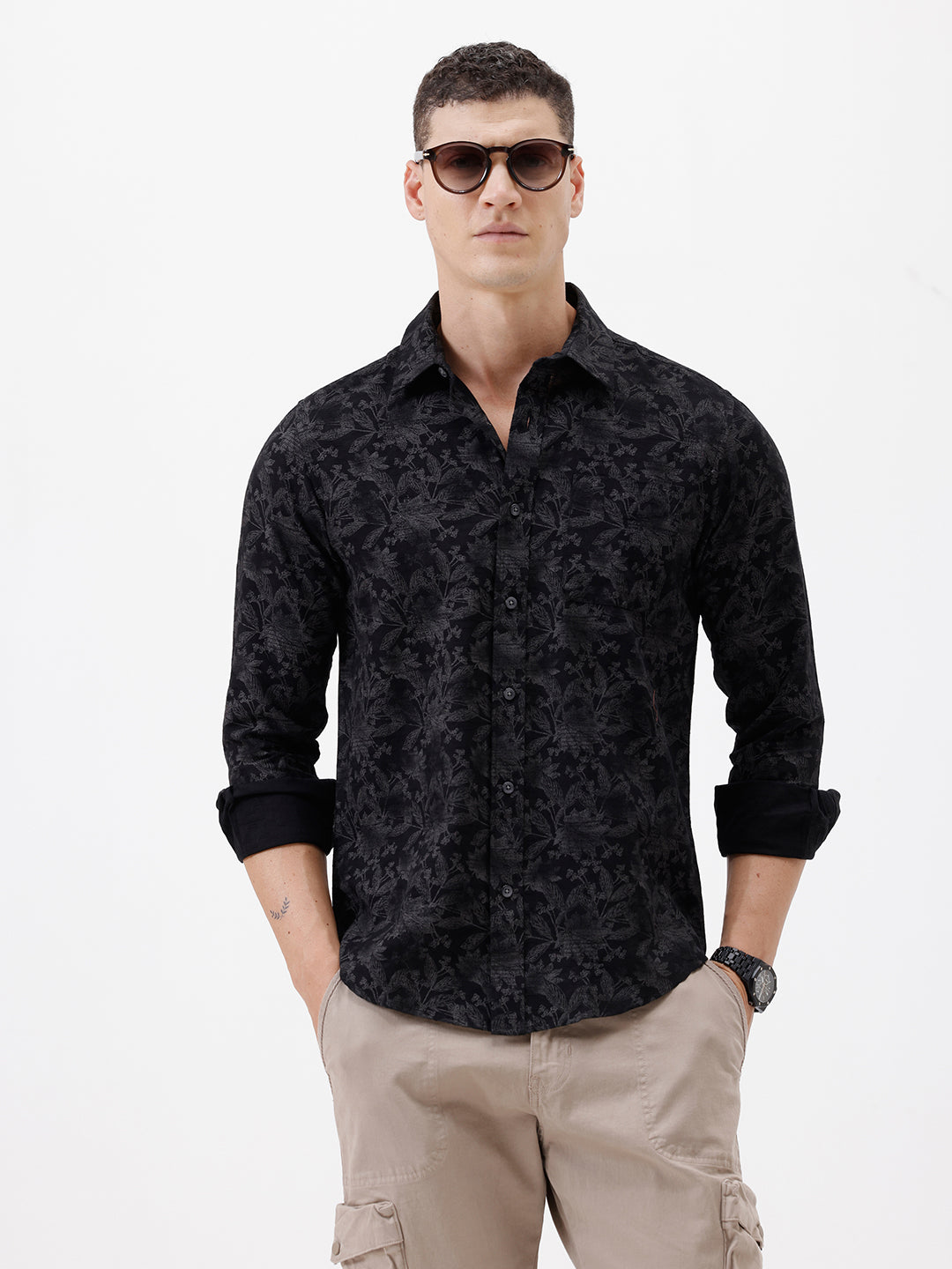 A man wearing a black long-sleeve button-up shirt with a subtle tonal floral pattern, paired with beige cargo pants and dark sunglasses, standing against a plain white background.