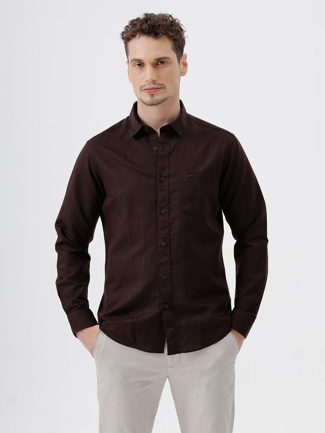 A man wearing a dark brown long-sleeve button-down shirt and light beige trousers, standing against a plain light gray studio background.