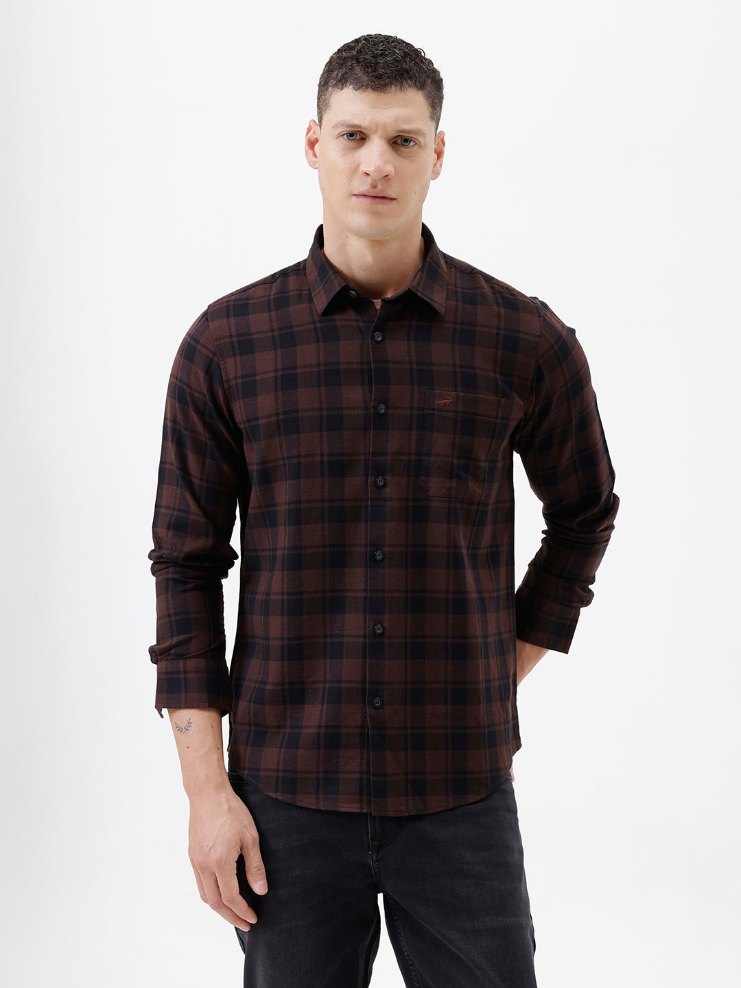 A man wearing a dark brown and black plaid long-sleeve button-up shirt, paired with dark pants, standing against a plain light gray background.