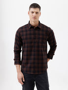 A man wearing a dark brown and black plaid long-sleeve button-up shirt, paired with dark pants, standing against a plain light gray background.