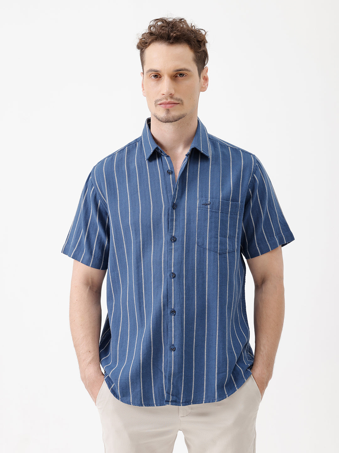 A man wearing a blue short-sleeve button-up shirt with thin white vertical stripes and beige trousers, standing against a plain white studio background.