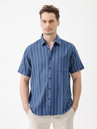 A man wearing a blue short-sleeve button-up shirt with thin white vertical stripes and beige trousers, standing against a plain white studio background.