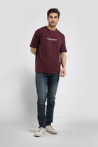Man wearing a maroon t-shirt with a logo and dark jeans on a white background