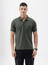A man wearing a dark olive green short-sleeve polo shirt with a chest pocket, paired with dark jeans, standing against a plain light gray background.