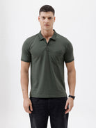 A man wearing a dark olive green short-sleeve polo shirt with a chest pocket, paired with dark jeans, standing against a plain light gray background.