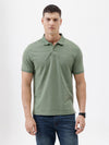 A man wearing a short-sleeve polo shirt in sage green, paired with dark blue jeans, standing against a plain light gray background.