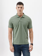 A man wearing a short-sleeve polo shirt in sage green, paired with dark blue jeans, standing against a plain light gray background.