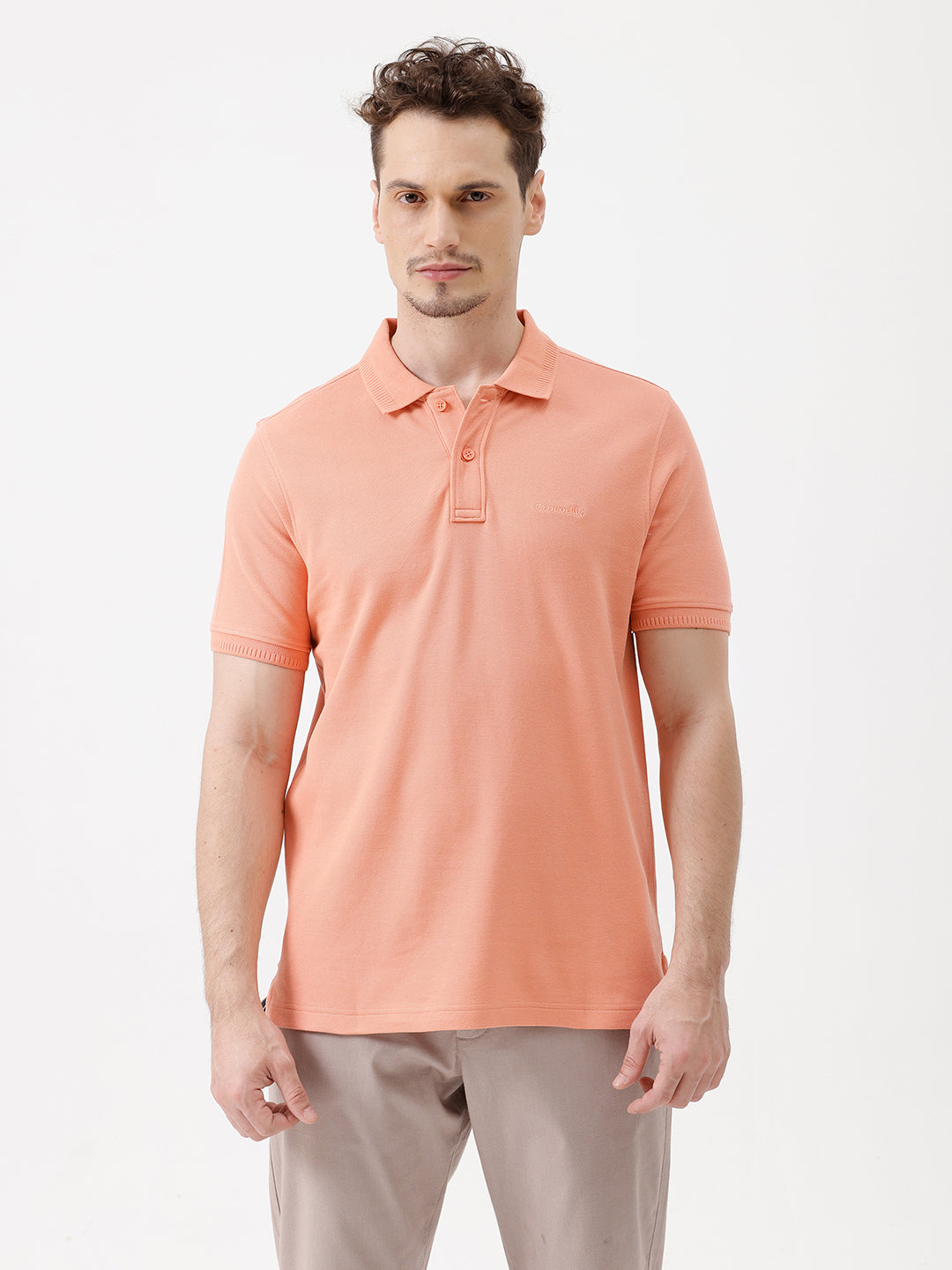 A man wearing a peach-colored short-sleeve polo shirt and beige trousers, standing against a plain white studio background.