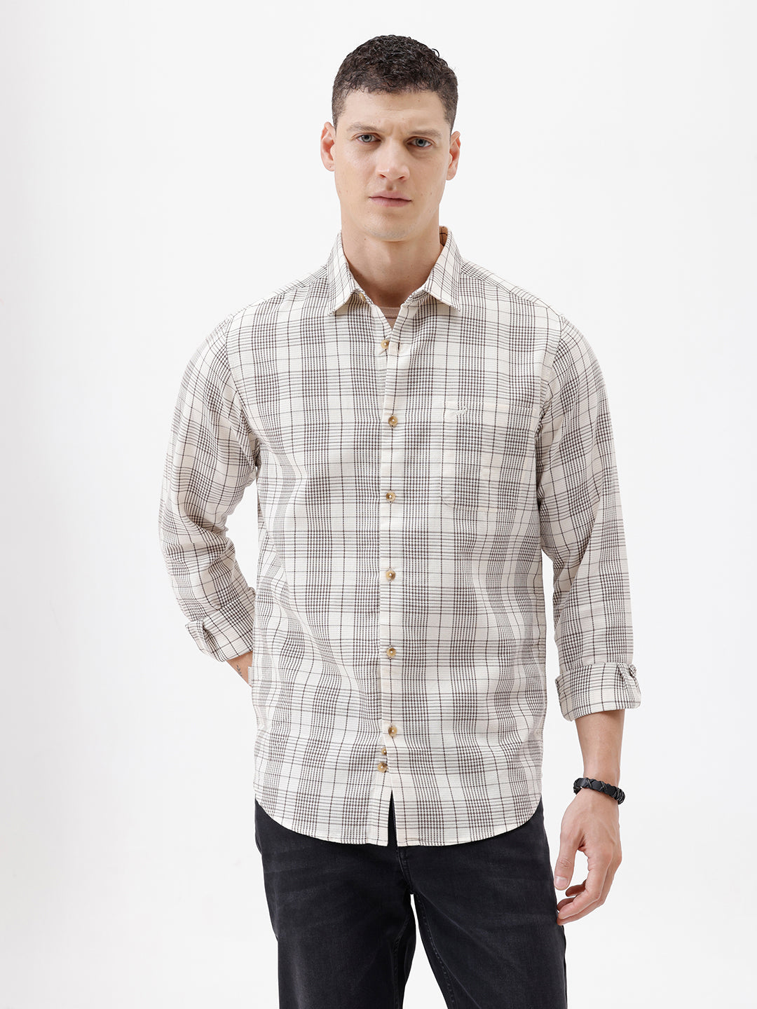 A man wearing a long-sleeve button-up plaid shirt in beige, white, and gray, paired with dark pants, standing against a plain white studio background.