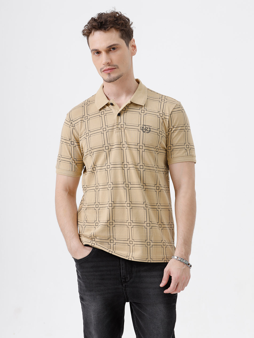 A man wearing a beige short-sleeve polo shirt with a brown grid pattern, paired with black jeans, standing against a plain white studio background.