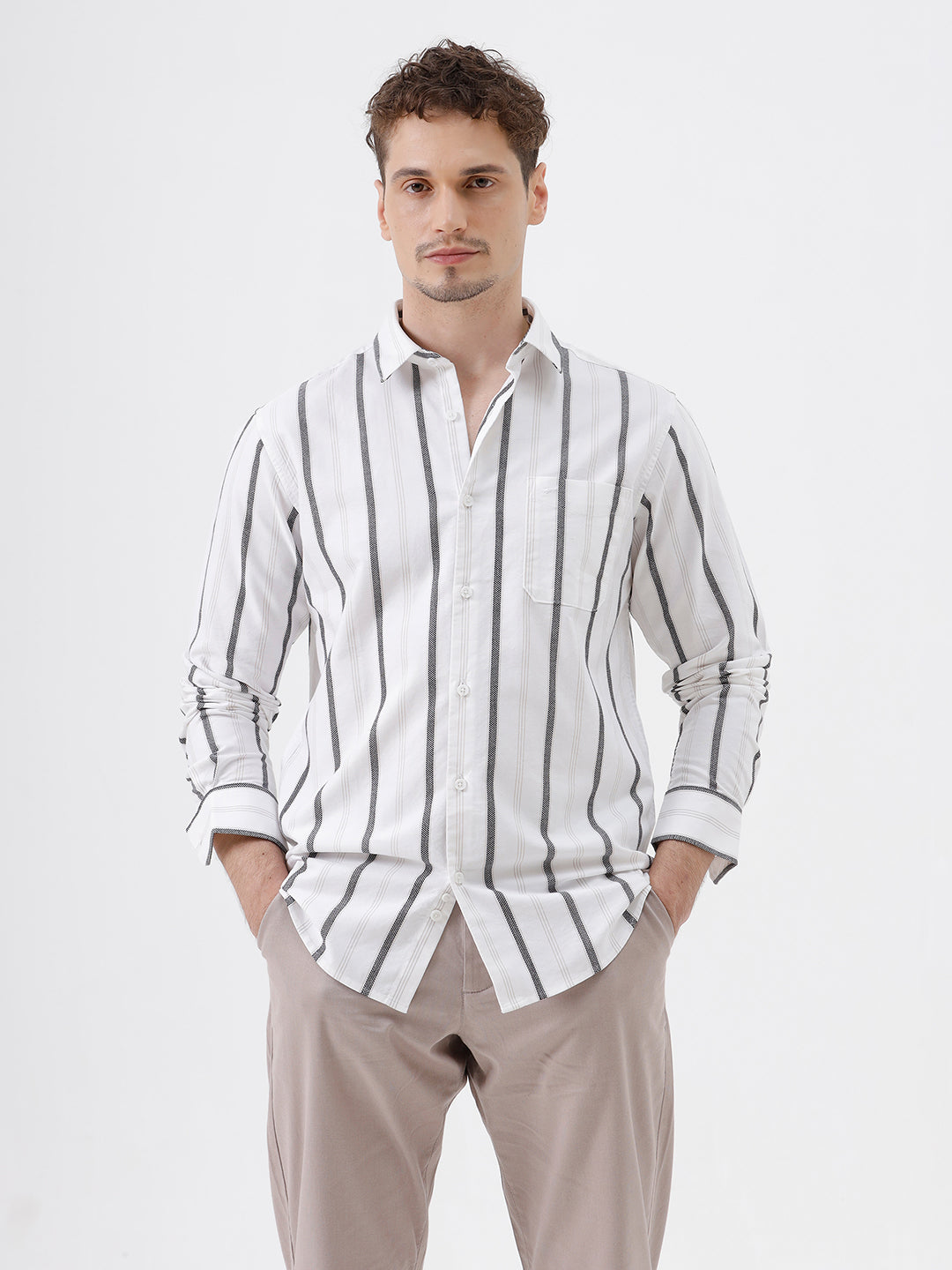 A man wearing a white long-sleeve button-up shirt with vertical gray stripes and beige trousers, standing against a light gray studio background.
