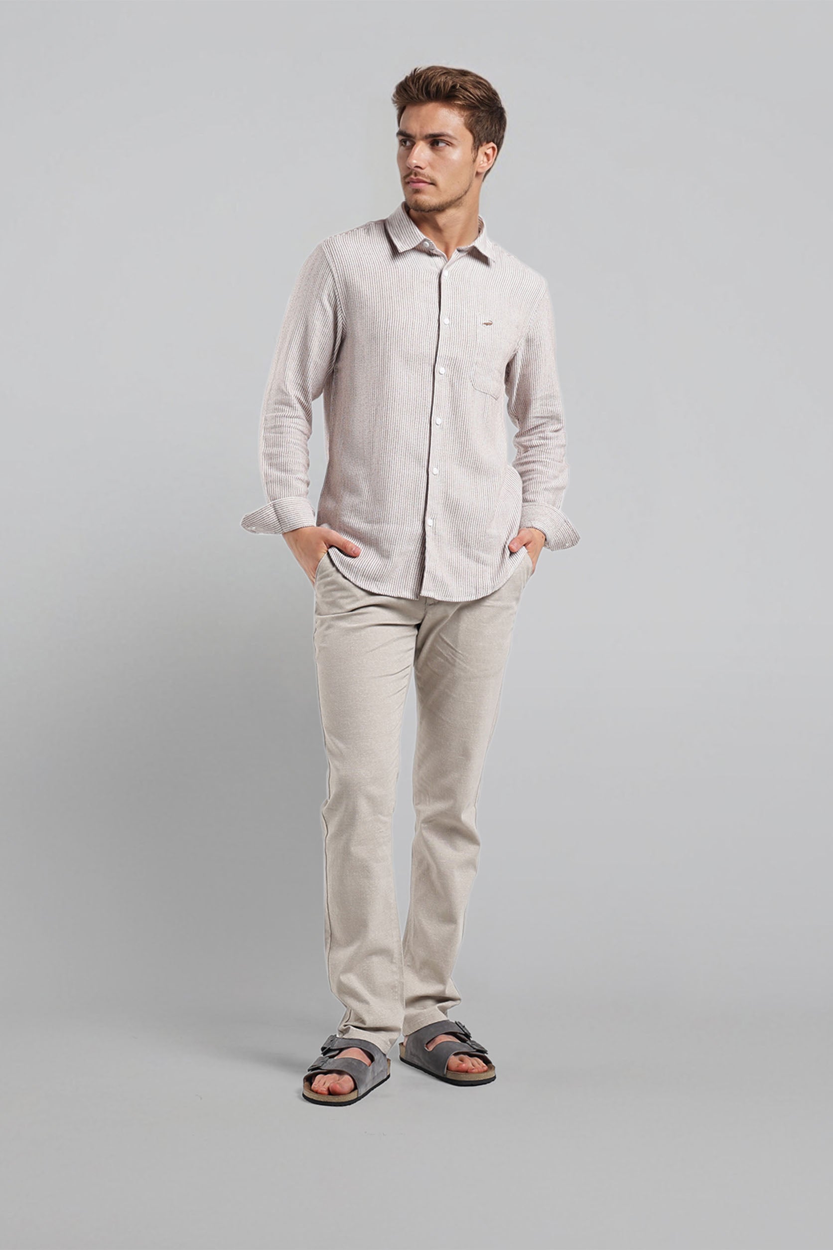Man wearing a light gray shirt and pants with sandals on a plain background