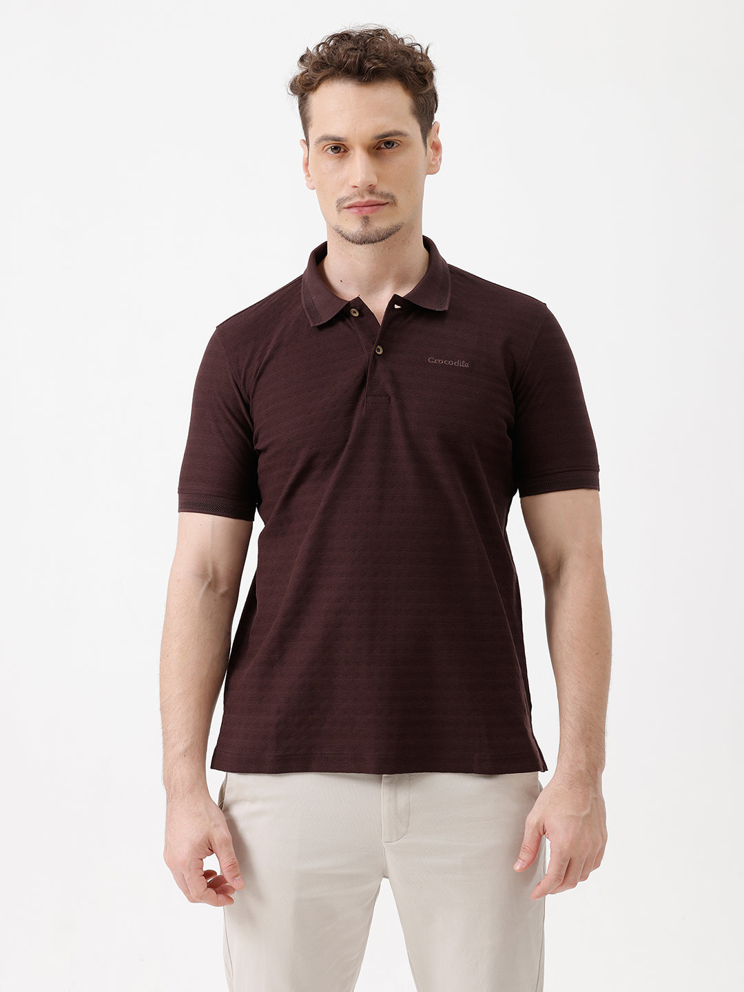 A man wearing a dark brown short-sleeve polo shirt with a subtle horizontal stripe pattern and light beige trousers, standing against a plain white studio background.
