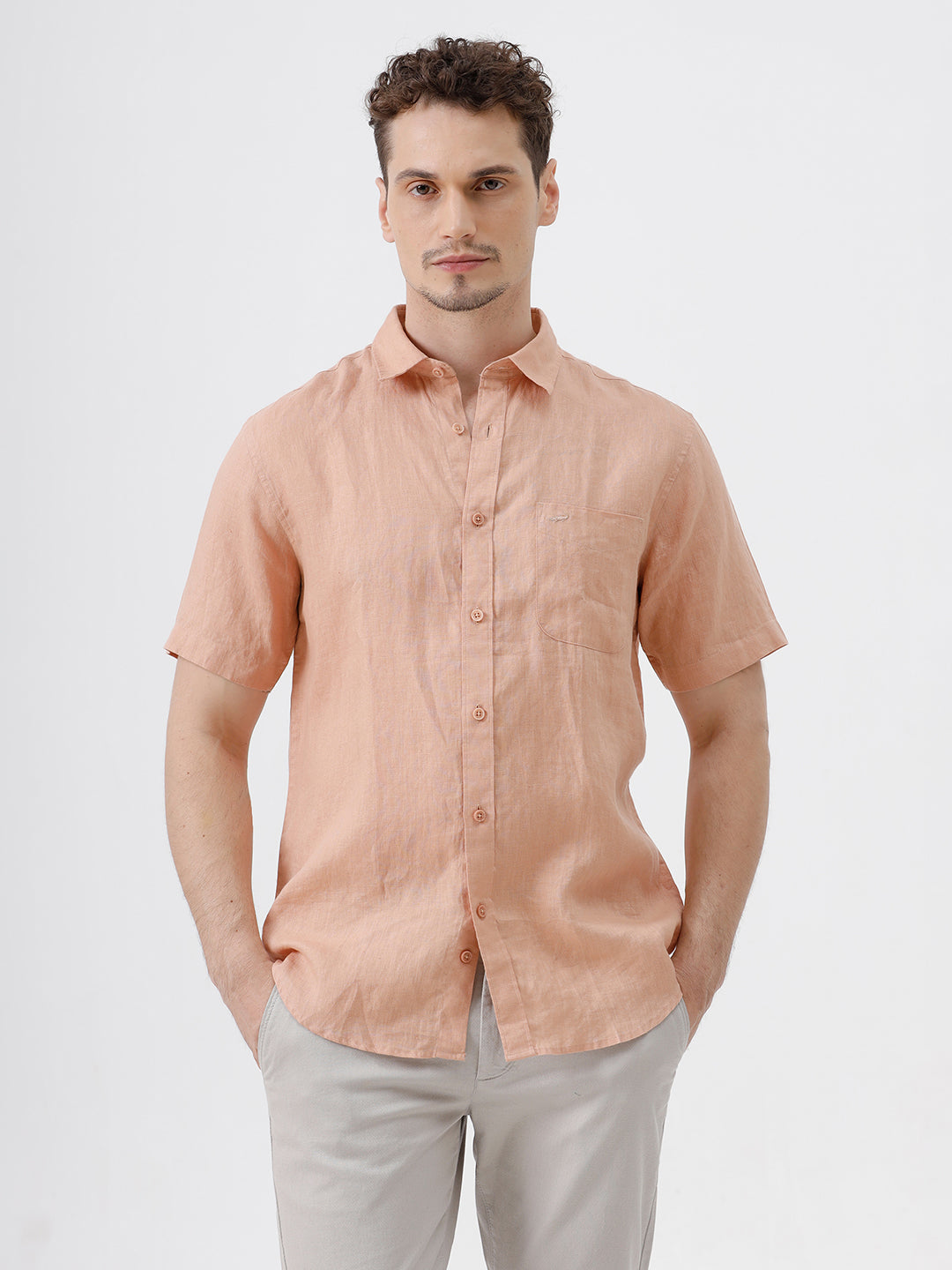 A man wearing a short-sleeve linen button-up shirt in a light peach-pink color, paired with light gray trousers, standing against a plain light gray background.