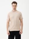 A man wearing a light beige short-sleeve t-shirt with a subtle tonal logo on the chest, paired with black pants, standing against a plain white studio background.