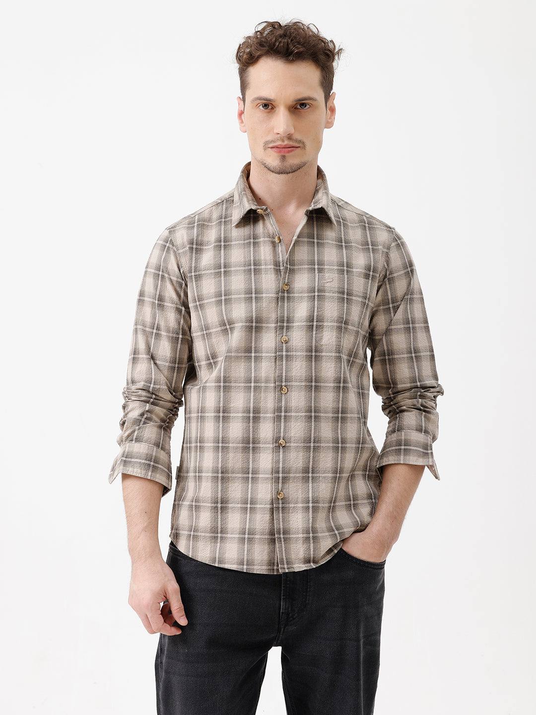 A man wearing a long-sleeve button-up plaid shirt in beige and brown tones, paired with dark jeans, standing against a plain white studio background.