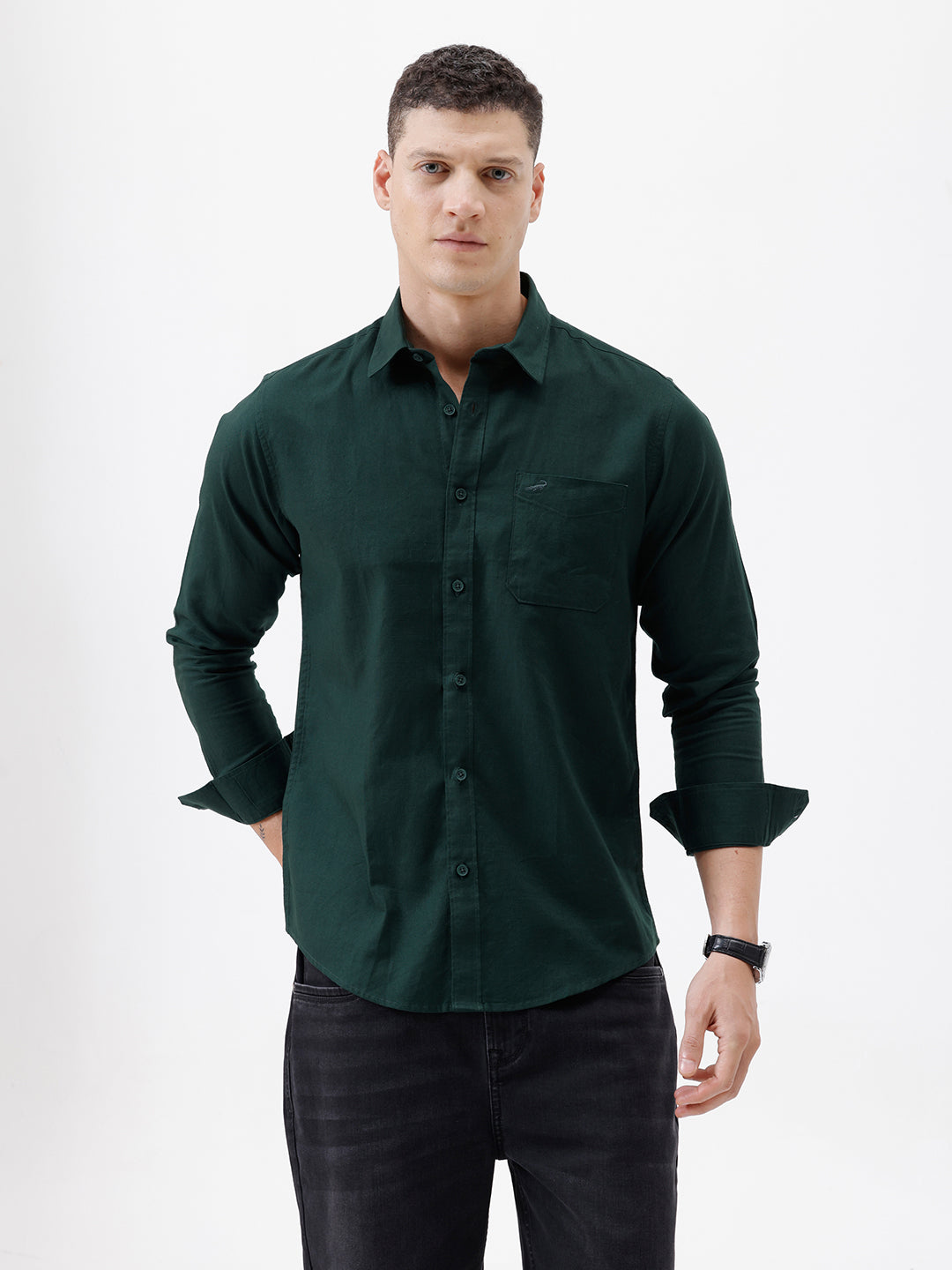 A man wearing a dark forest green long-sleeve button-down shirt with a chest pocket, paired with dark jeans, standing against a plain white studio background.