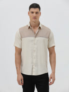 A man wearing a short-sleeve linen button-up shirt with a color-blocked design in beige and cream, paired with black pants, against a plain white studio background.