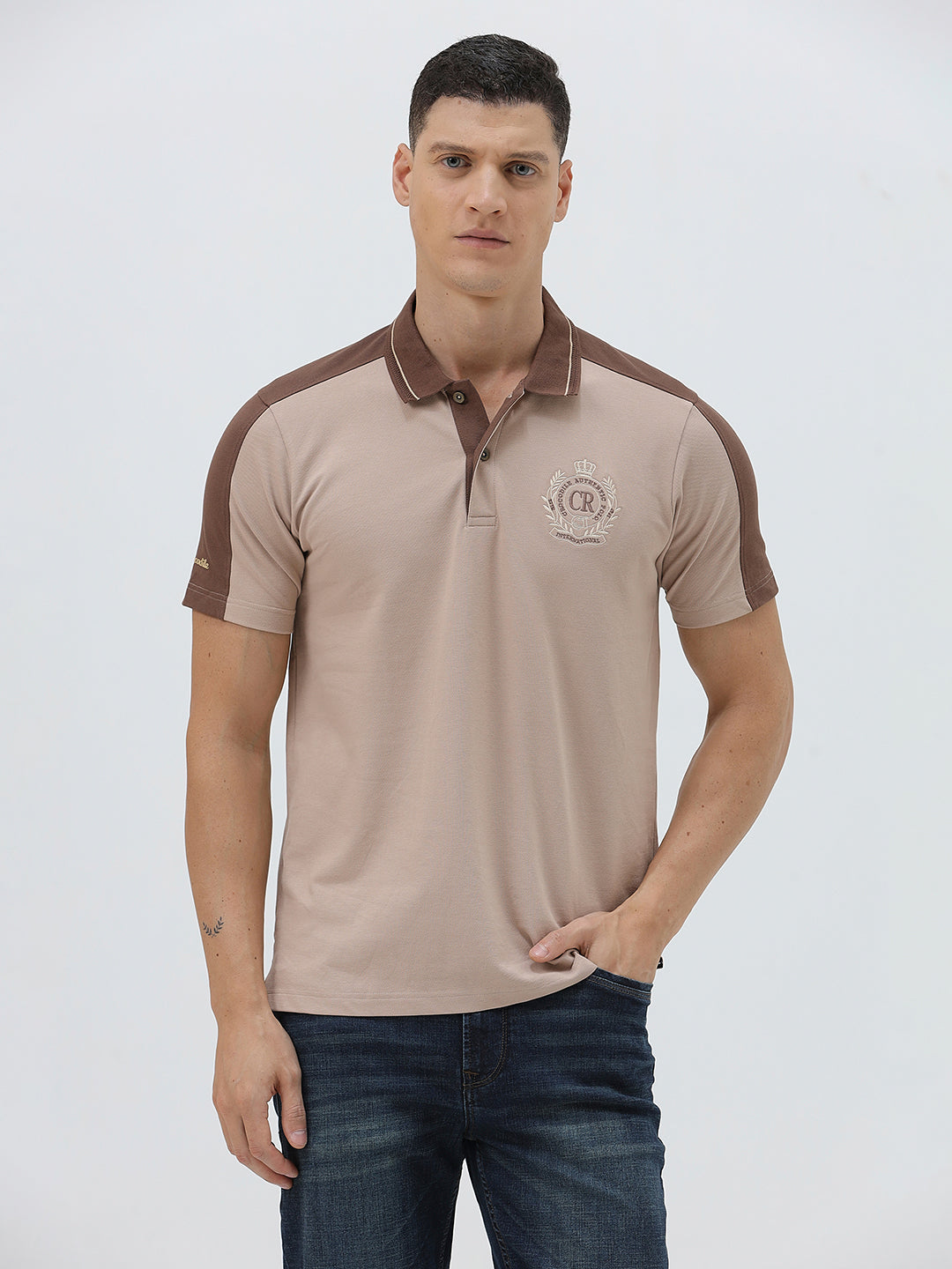 A man modeling a short-sleeve polo shirt in beige with brown shoulder panels and collar trim, paired with dark blue jeans, against a light gray studio background.