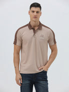 A man modeling a short-sleeve polo shirt in beige with brown shoulder panels and collar trim, paired with dark blue jeans, against a light gray studio background.