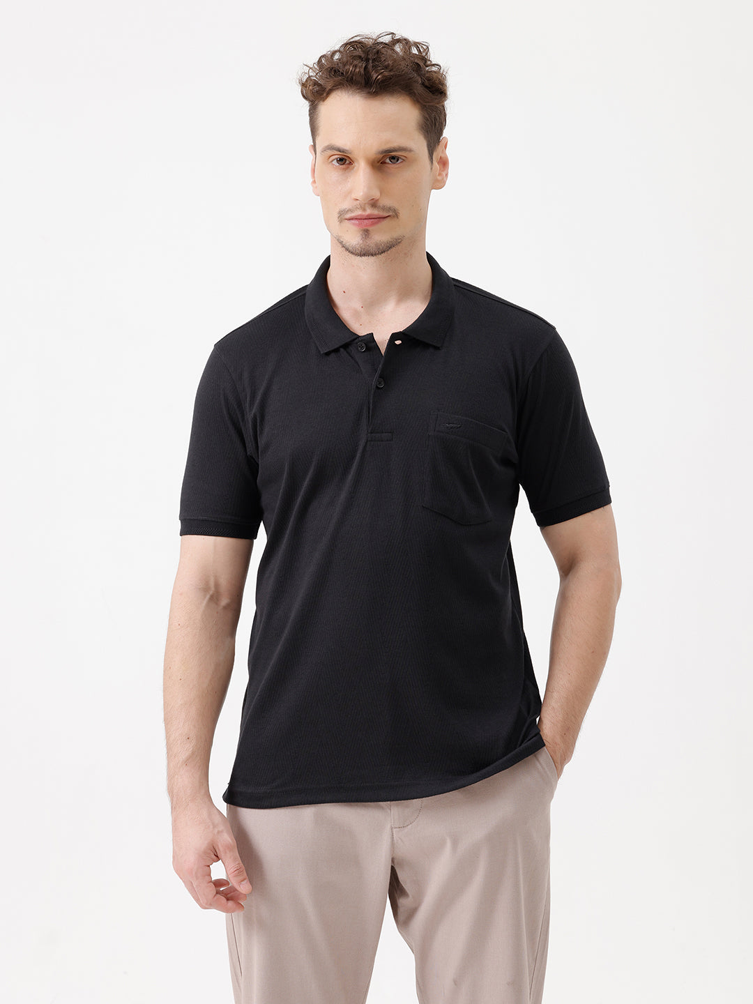 A man wearing a black short-sleeve polo shirt with a single chest pocket, paired with beige trousers, standing against a plain white studio background.