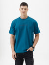 A man wearing a teal blue short-sleeve t-shirt with a subtle tonal 'GREATNESS' text graphic across the chest, paired with black pants, photographed against a clean white studio background.