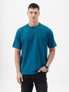A man wearing a teal blue short-sleeve t-shirt with a subtle tonal 'GREATNESS' text graphic across the chest, paired with black pants, photographed against a clean white studio background.