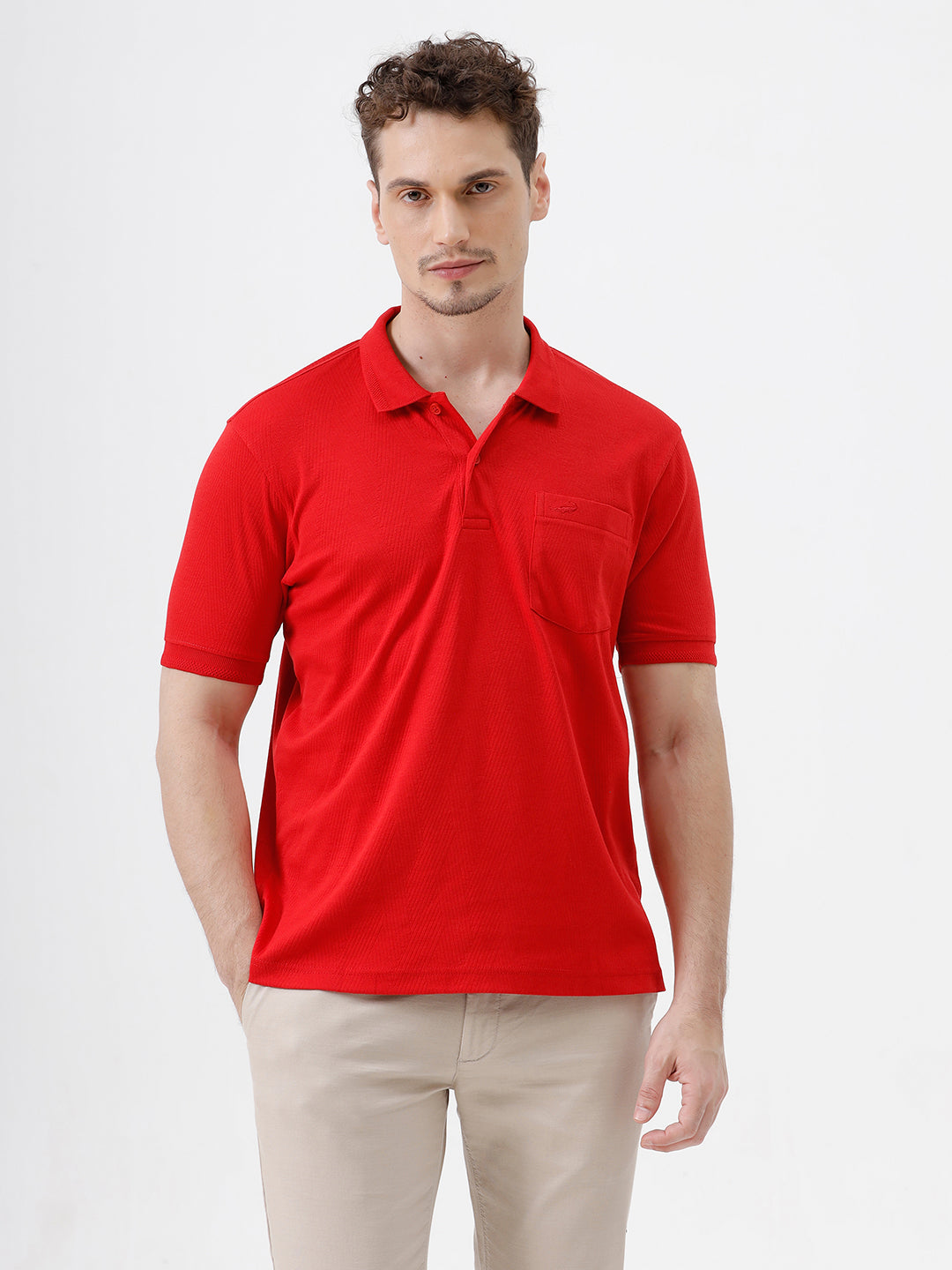 A man wearing a bright red short-sleeve polo shirt with a chest pocket, paired with beige trousers, standing against a plain white studio background.