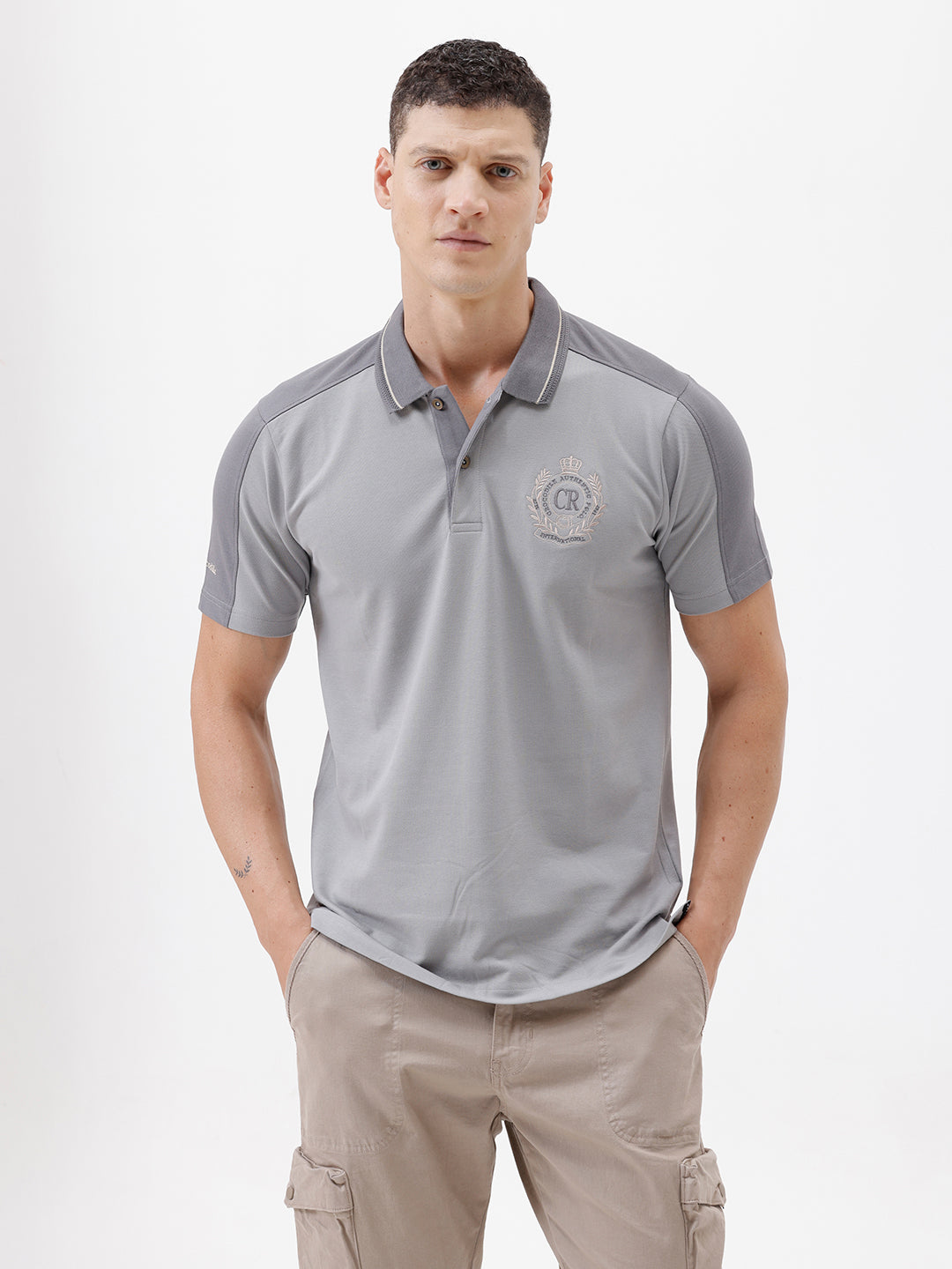 A man wearing a light gray short-sleeve polo shirt with darker gray shoulder panels and a small crest logo on the left chest, paired with khaki cargo pants, against a plain light gray studio background.