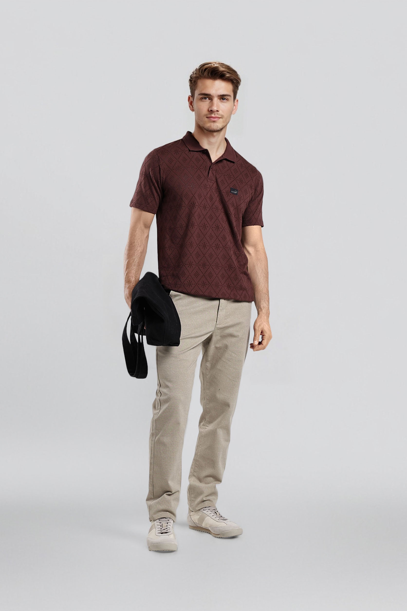 Man wearing a maroon polo shirt and beige pants holding a black jacket on a light gray background