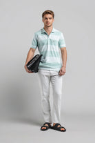 Man wearing a light blue striped polo shirt and white pants, holding a black bag on a gray background