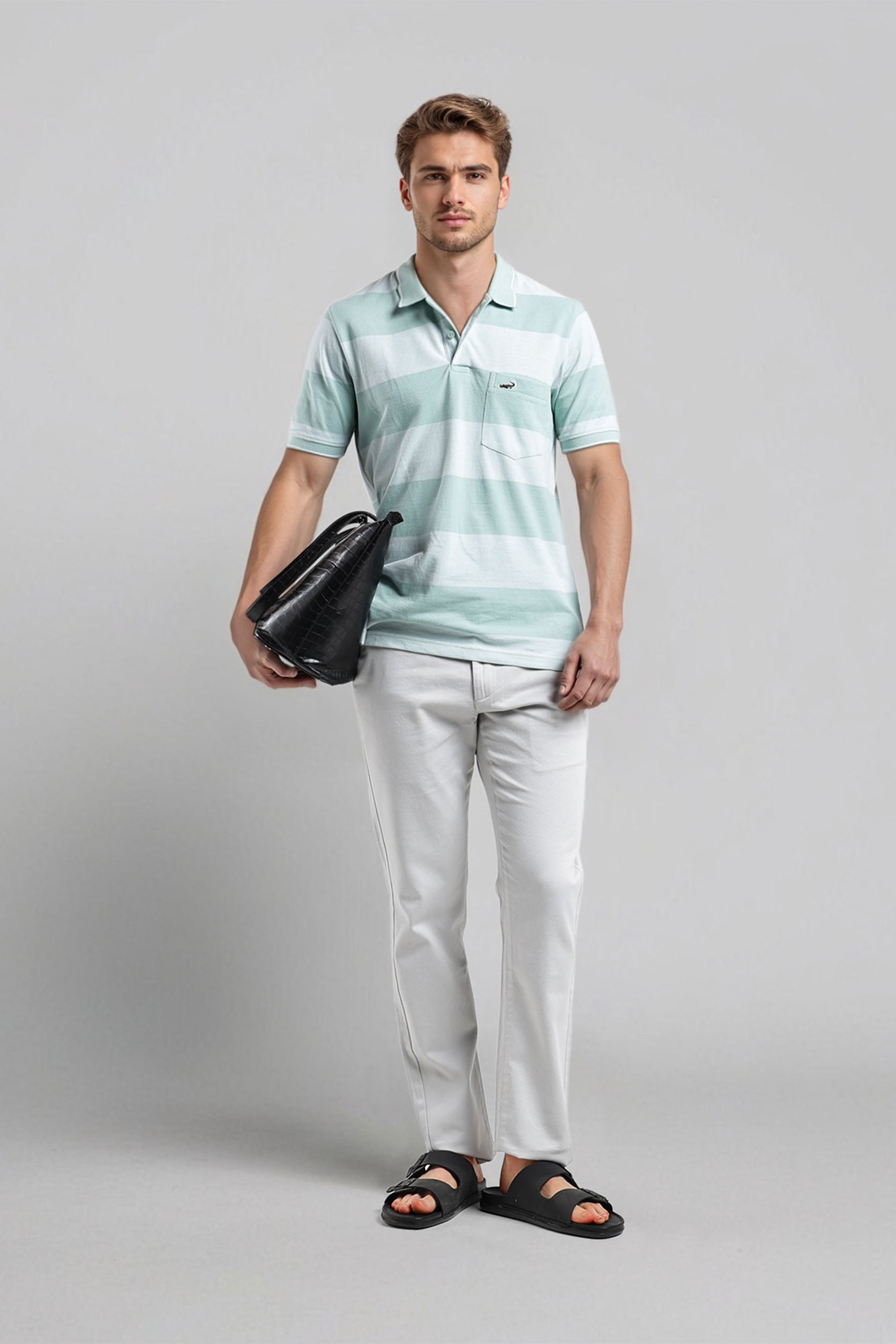 Man wearing a light blue striped polo shirt and white pants, holding a black bag on a gray background