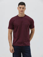A man wearing a short-sleeve crewneck t-shirt in maroon with a subtle tonal graphic pattern, paired with dark blue jeans, against a light gray studio background.