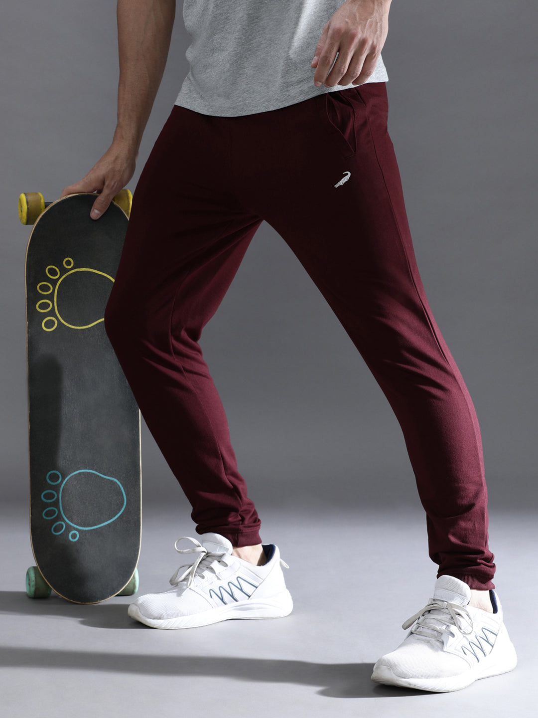 A person wearing wine red solid joggers with a comfort fit, standing next to a skateboard, wearing a grey top and white sneakers.