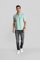 Man wearing a light green patterned polo shirt and dark gray jeans on a gray background