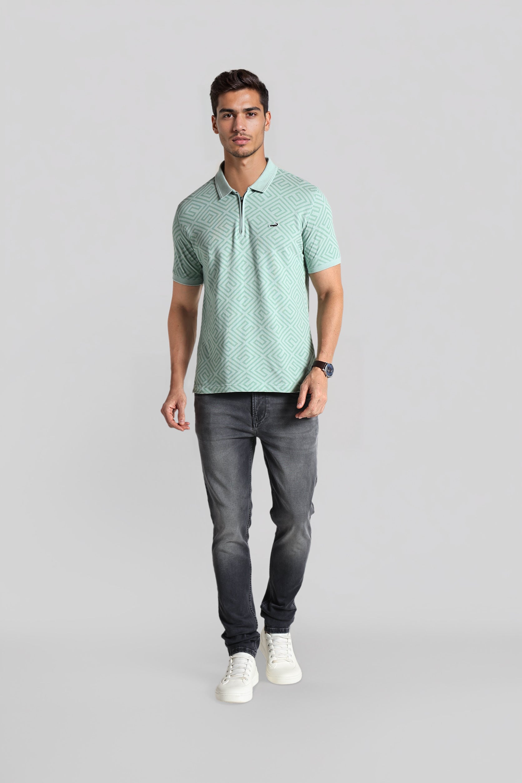 Man wearing a light green patterned polo shirt and dark gray jeans on a gray background