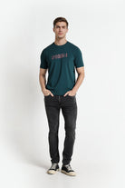 Man wearing a green t-shirt with text and dark jeans on a white background