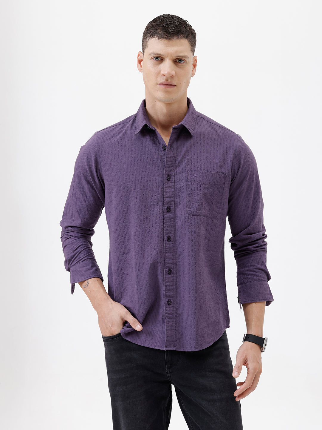 A man wearing a long-sleeve button-down shirt in a muted purple color, paired with black pants, standing against a plain white studio background.