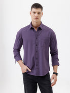 A man wearing a long-sleeve button-down shirt in a muted purple color, paired with black pants, standing against a plain white studio background.