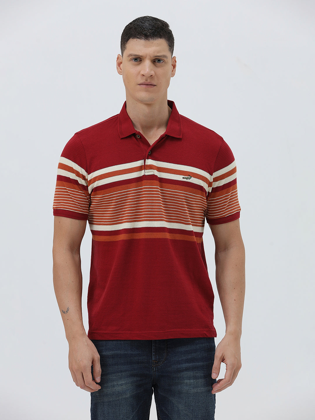 A man wearing a red short-sleeve polo shirt with horizontal cream and rust-orange stripes across the chest and sleeves, paired with dark blue jeans and black sunglasses, against a light gray studio background.