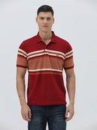 A man wearing a red short-sleeve polo shirt with horizontal cream and rust-orange stripes across the chest and sleeves, paired with dark blue jeans and black sunglasses, against a light gray studio background.