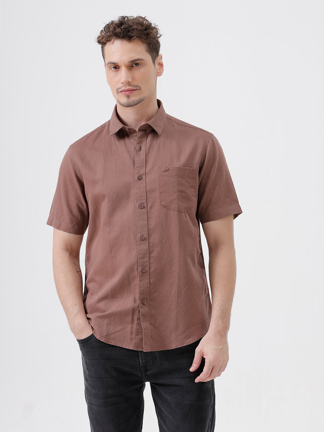 A man wearing a short-sleeve button-up shirt in a dusty rose-brown color, paired with black jeans, standing against a plain light gray background.