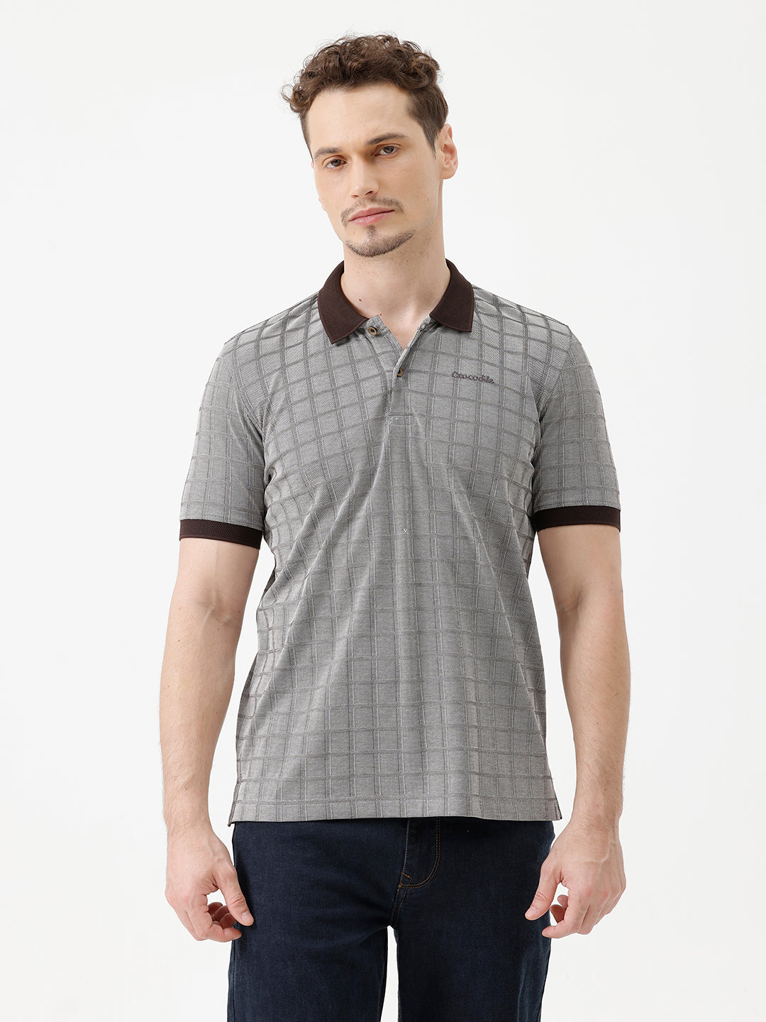 A man wearing a gray short-sleeve polo shirt with a subtle grid pattern and dark brown collar and sleeve trim, paired with dark blue jeans, against a plain white studio background.