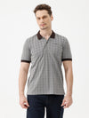 A man wearing a gray short-sleeve polo shirt with a subtle grid pattern and dark brown collar and sleeve trim, paired with dark blue jeans, against a plain white studio background.