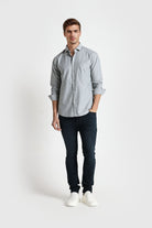 Man wearing a light gray button-up shirt and dark jeans on a white background
