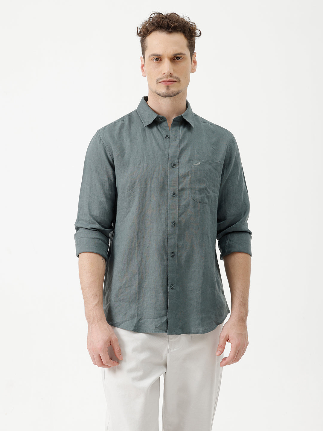 A man wearing a slate blue-green long-sleeve linen button-up shirt with white trousers, standing against a plain white studio background.