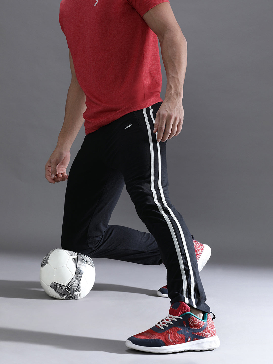 Person in red shirt and black pants with white stripes, kicking a soccer ball on a gray background