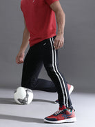 Person in red shirt and black pants with white stripes, kicking a soccer ball on a gray background