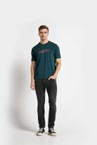 Man wearing a teal t-shirt with text and dark jeans on a white background